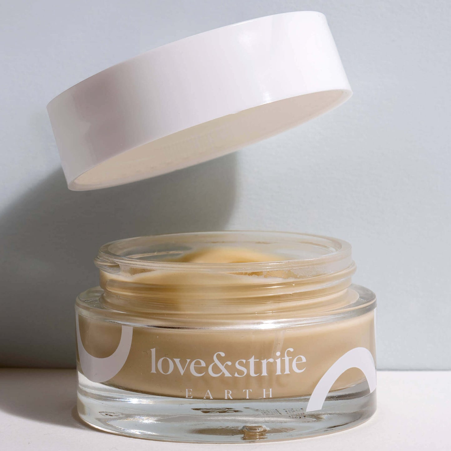 A glass jar of "love&strife earth" overnight mask with a white lid floating above it, partially open, against a plain light background.