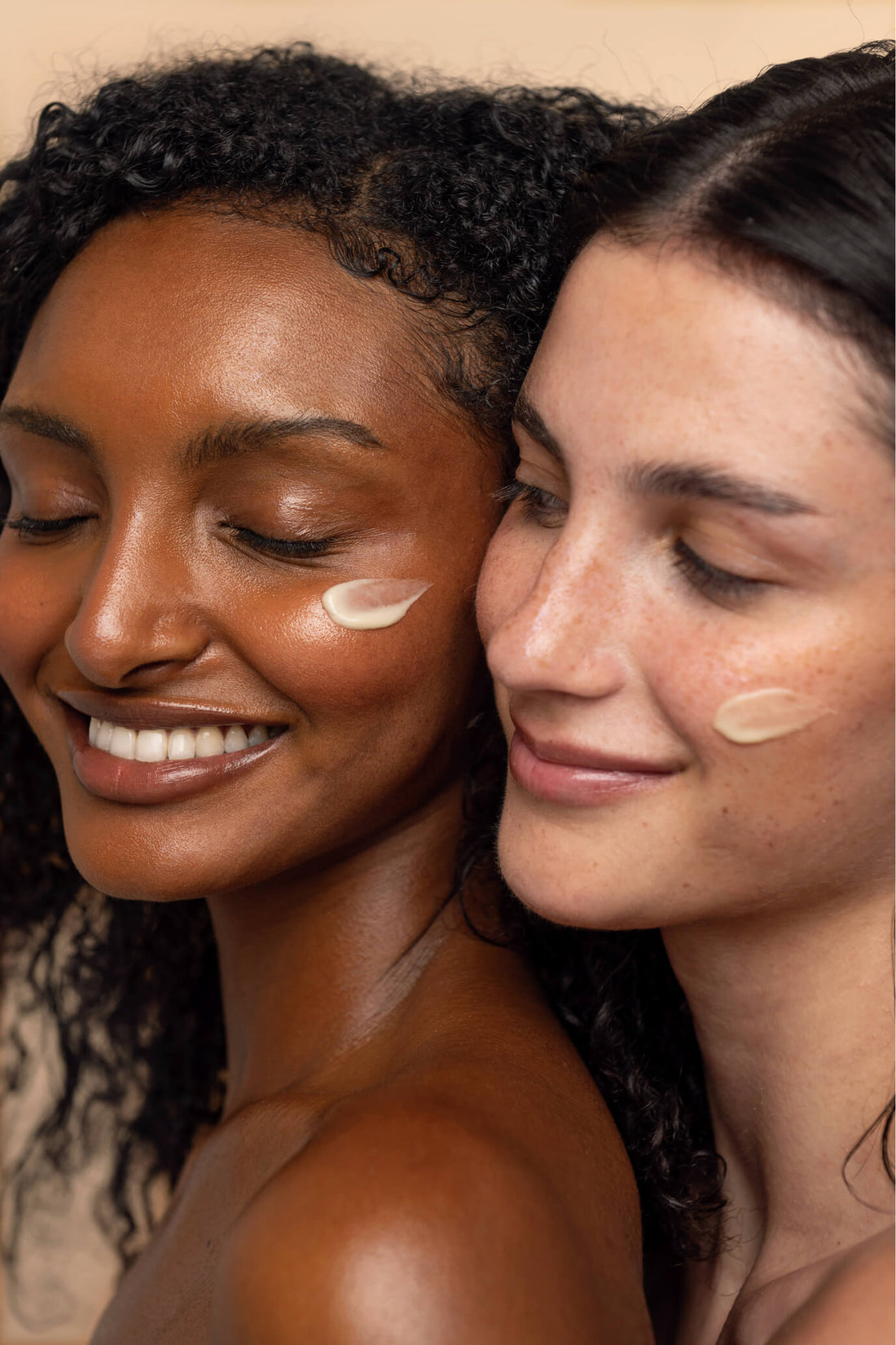 Two women models, one Caucasian and one Black, standing one behind the other with face cream applied on their cheeks.