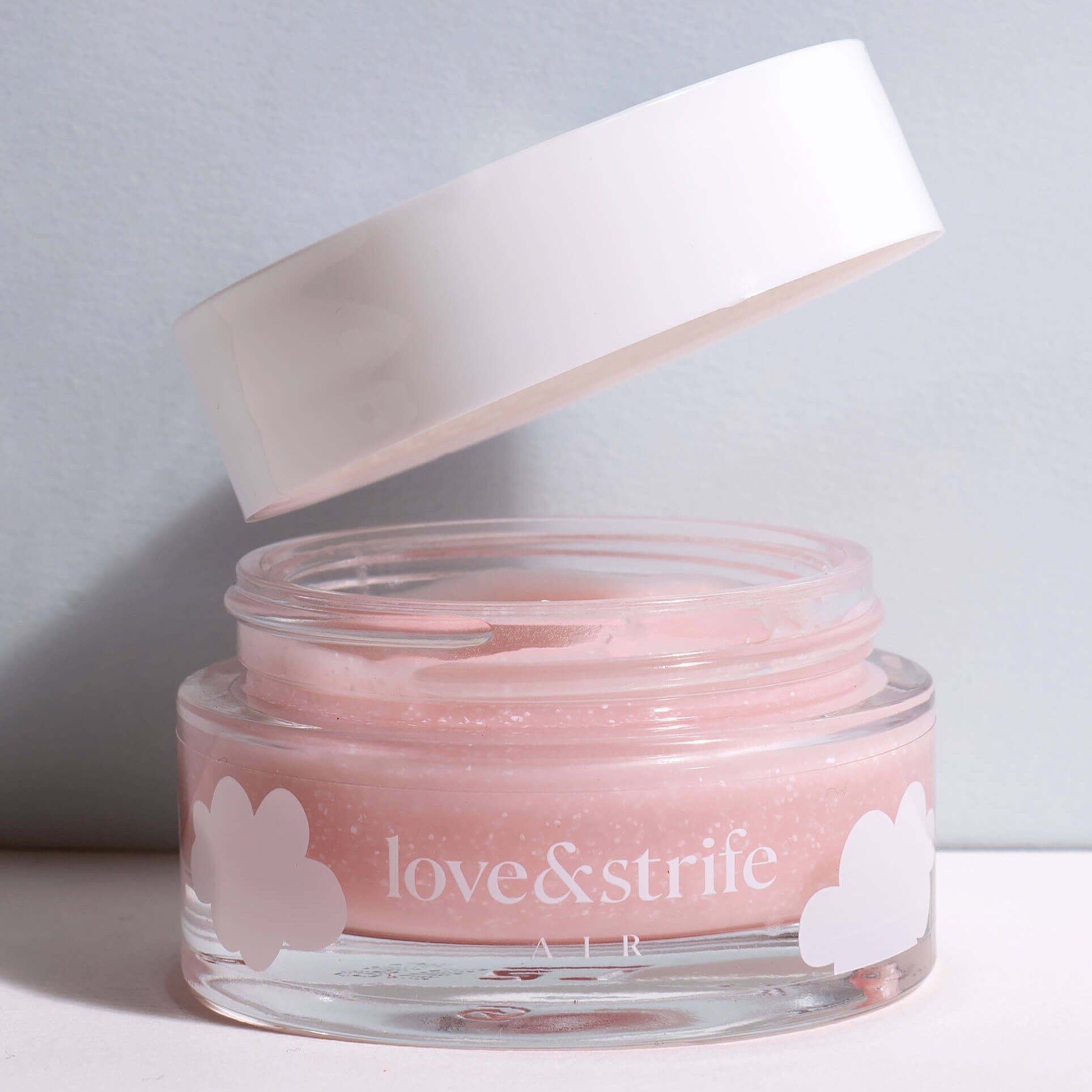 A glass jar of "love&strife air" exfoliating mask with a white lid floating above it, partially open, against a plain light background.