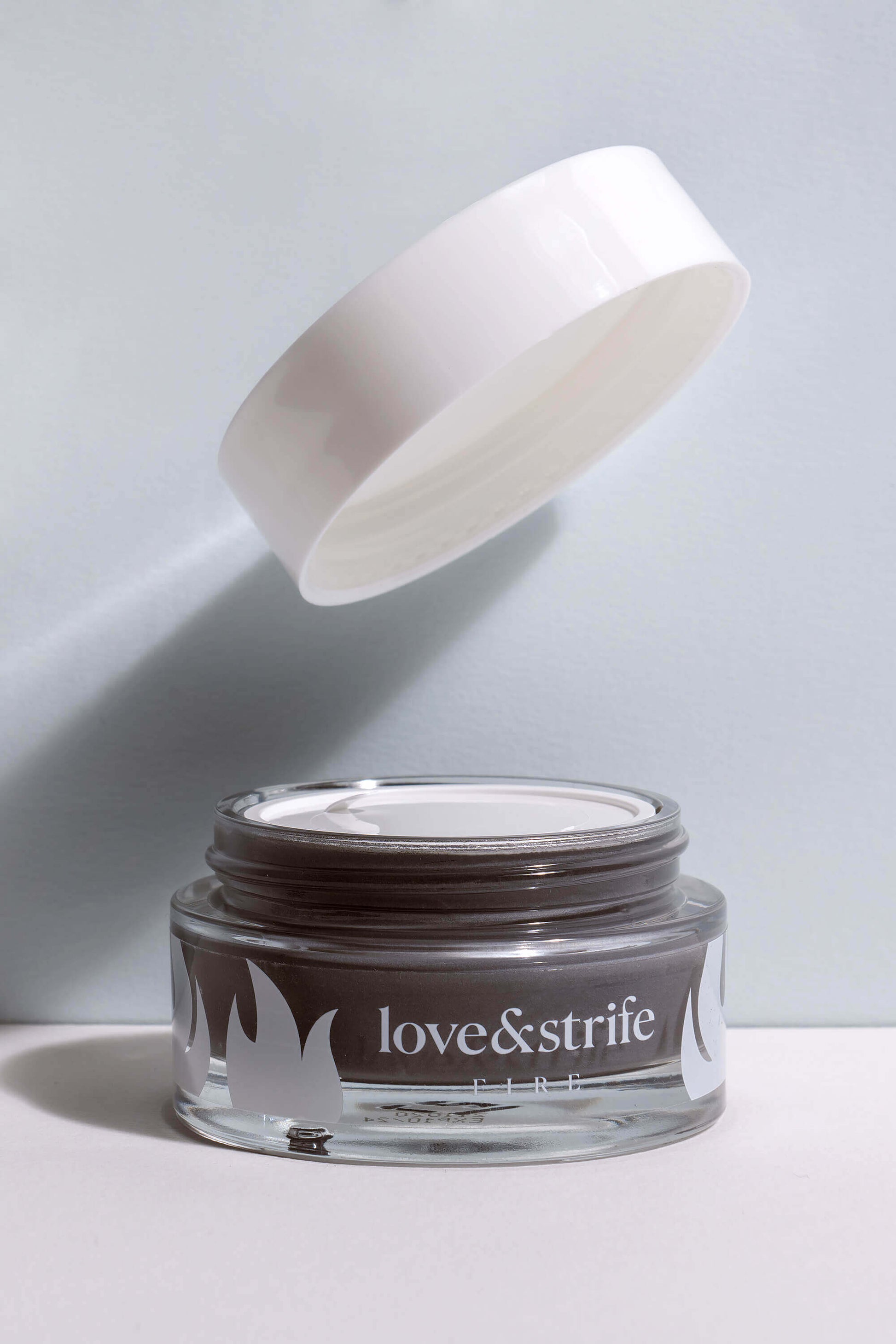 A glass jar of "love&strife fire" clay mask with a white lid floating above it, partially open, against a plain light background.