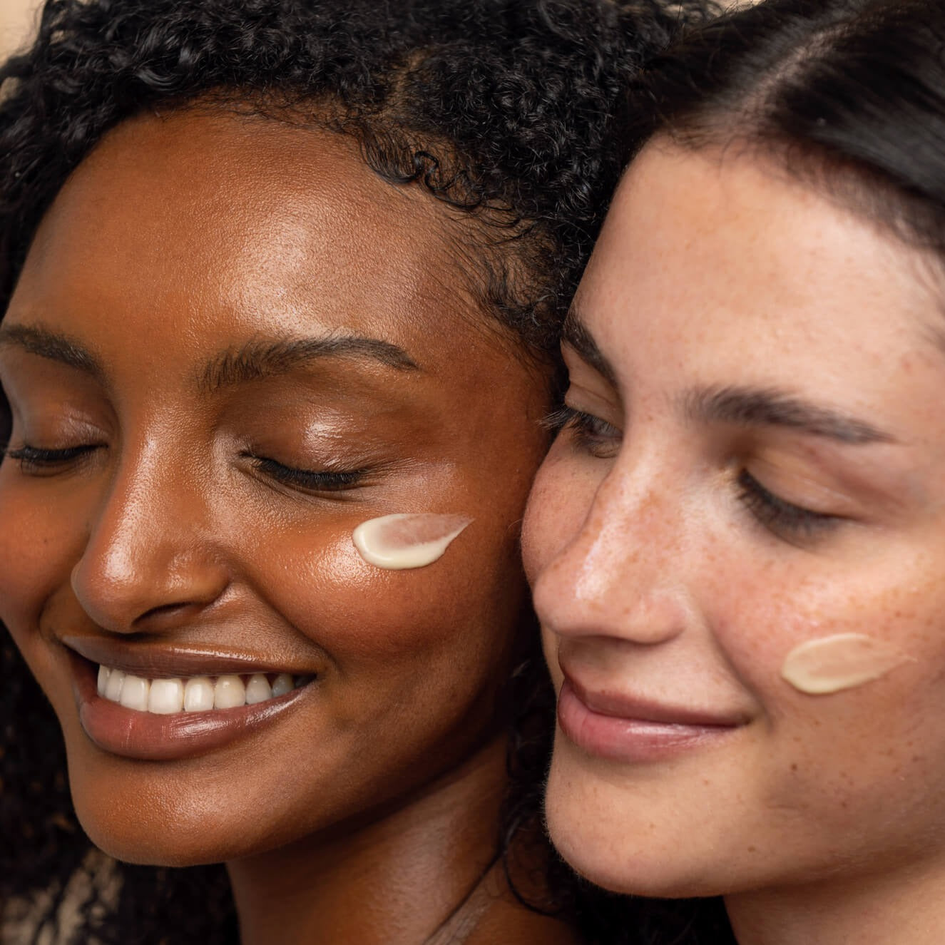 Two women models, one Caucasian and one Black, standing one behind the other with face cream applied on their cheeks.