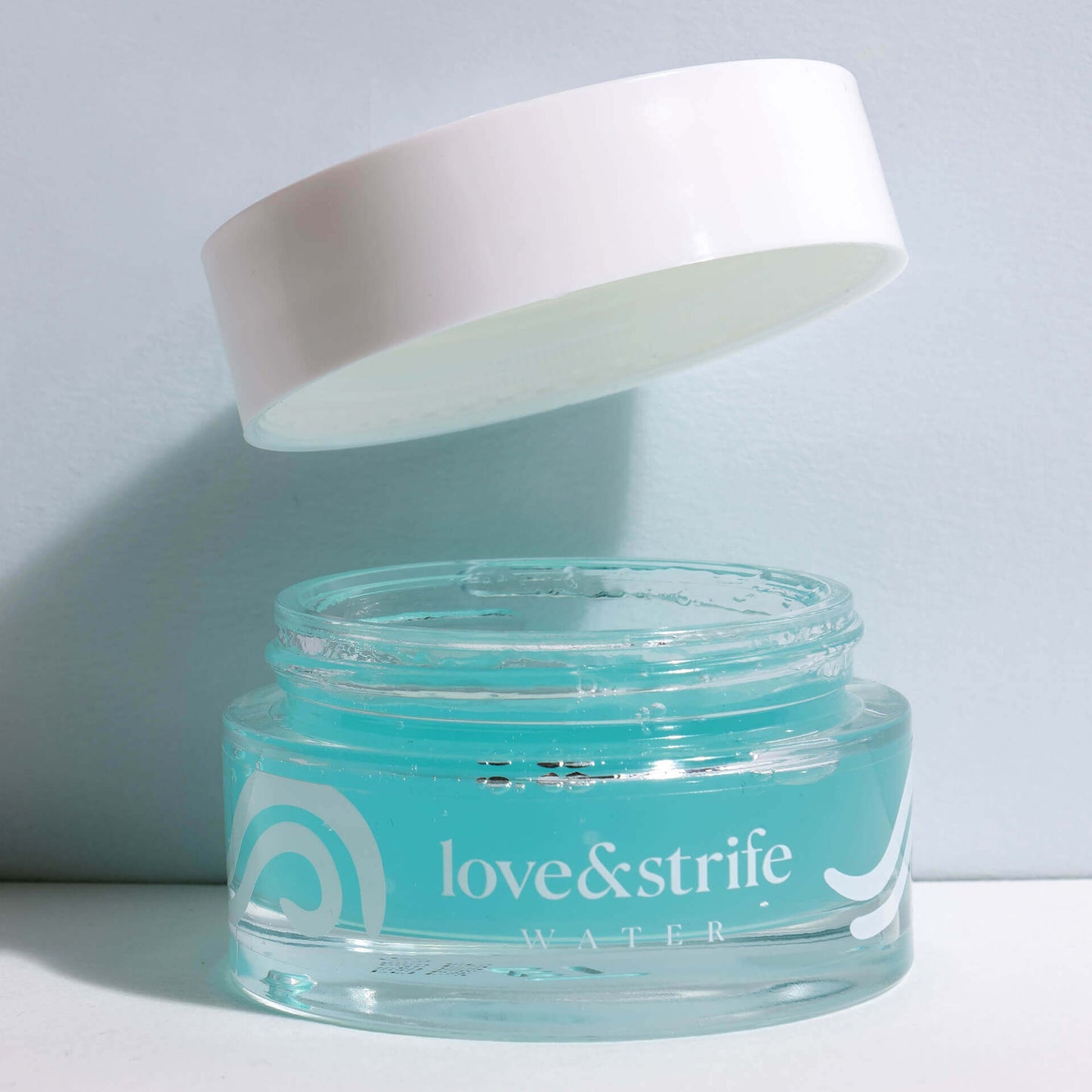 A glass jar of "love&strife water" gel mask with a white lid floating above it, partially open, against a plain light background.