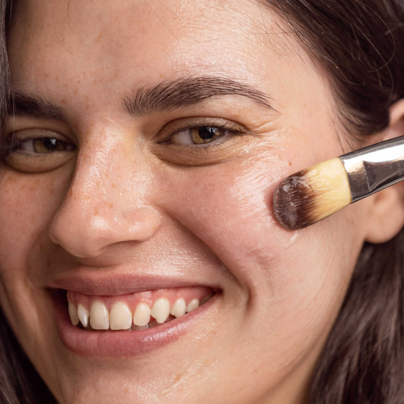 Young Caucasian woman looking directly at the camera smiling wide while applying a mask on her cheek with a makeup brush.